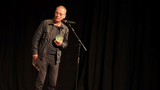 Thomas Heise steht am Mikrofon und hält den Preis (eine Schneekugel) in der rechten Hand und in der linken Hand die Urkunde