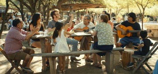 Menschen sitzen familiär in der Sonne an einem langem Tisch, mehrere Männer haben eine Gitarre