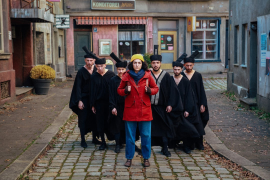 filmstill, eine frau mit roter jacke und gelber mütze, hinter ihr fünf männer und eine als mann verkleidete frau in traditionellen schwarzen gewändern