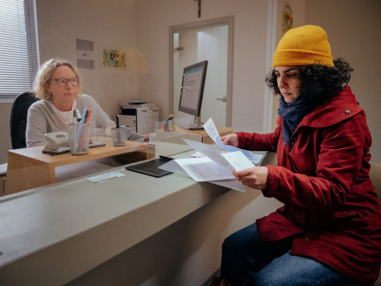 filmstill, narges sitzt im kreisverwaltungsamt, vor ihr durch eine glasscheibe getrennt die sachbearbeiterin, narges schaut sich formulare an